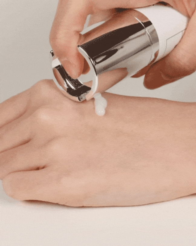 Hand dispensing white cream from silver and white pump bottle onto back of hand on white surface