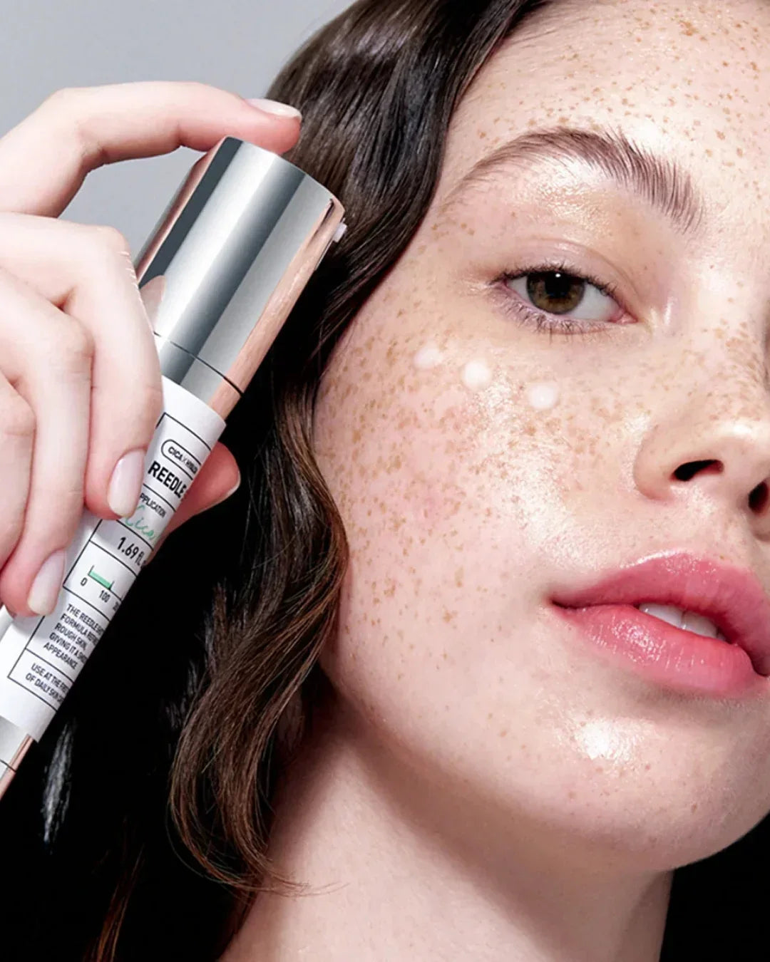 Close-up of freckled woman applying Reedle skincare serum with silver cap and white label