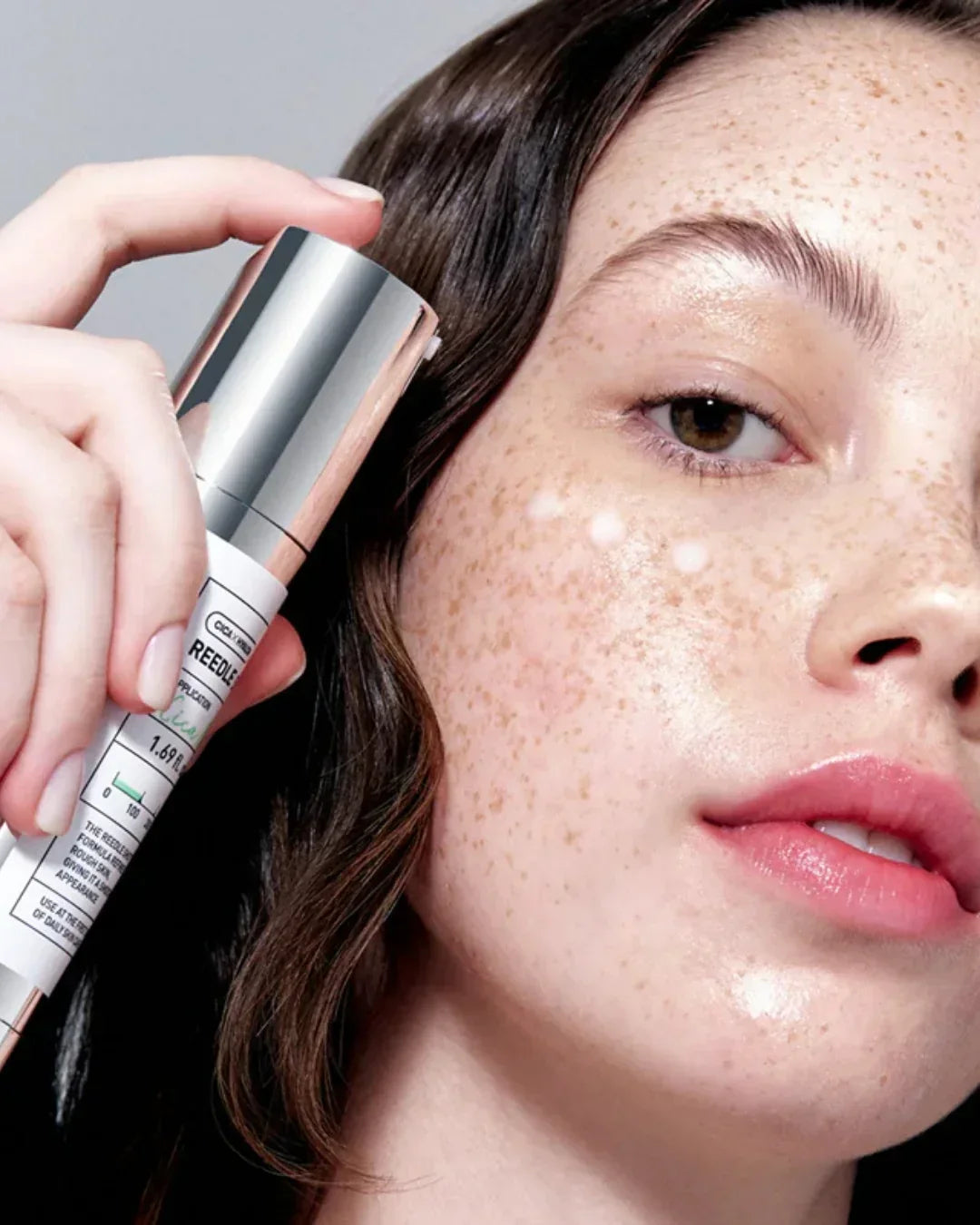 Close-up of freckled woman applying REEDLE skin serum with silver cap on cheek