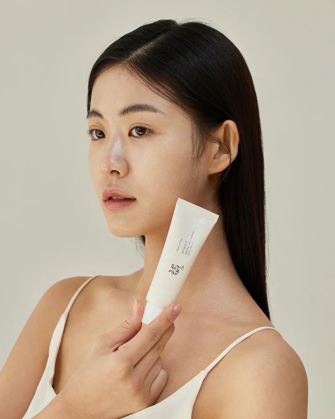 Young Asian woman with clear skin holding a white skincare tube in beige top against neutral background