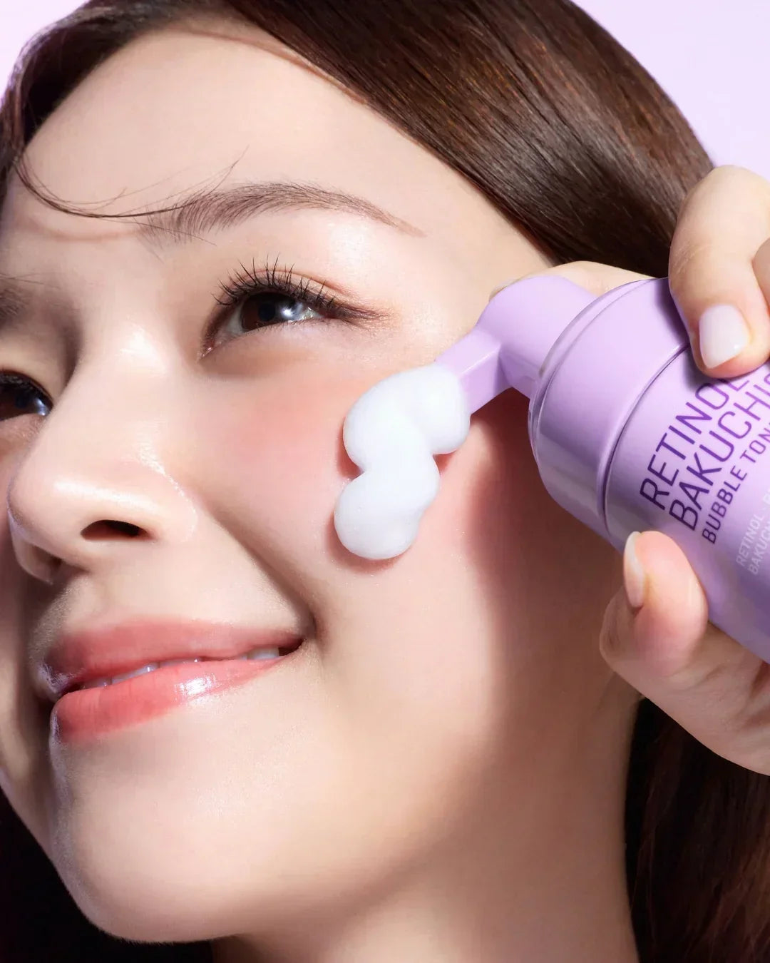 Close-up of woman applying Retinol Bakuchiol Bubble Toner foam on cheek with purple bottle