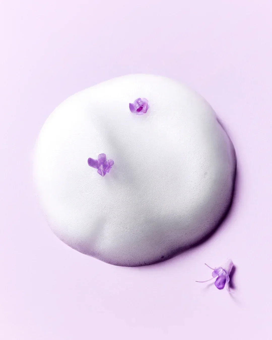 Foamy white skincare toner texture with small purple flower petals on a soft lavender background