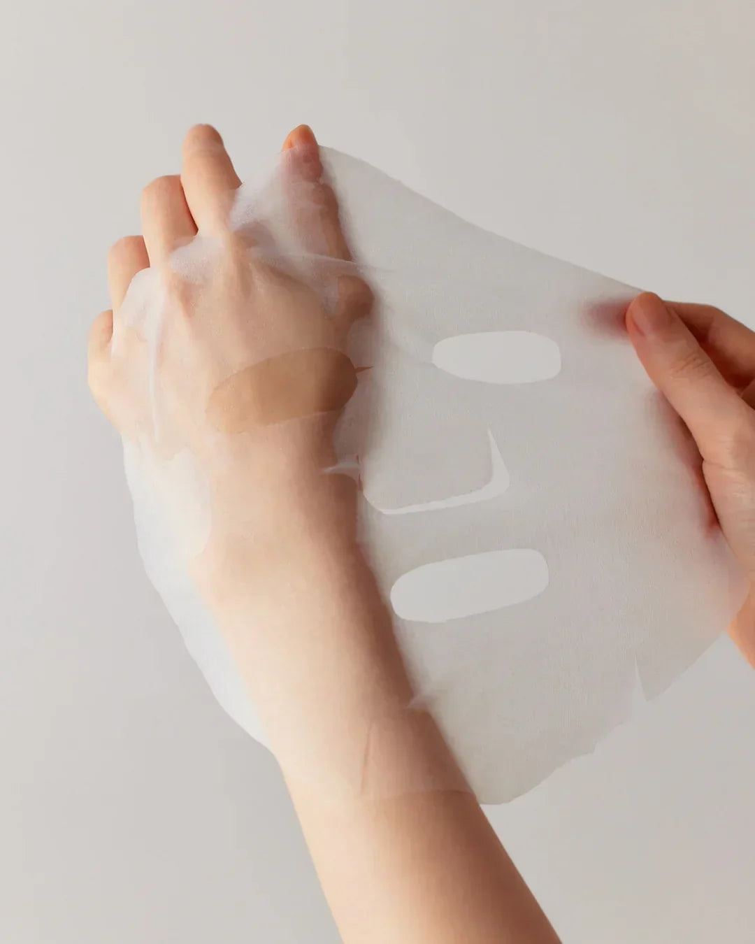 Person holding a thin white sheet face mask with cutouts for eyes and mouth against a plain background