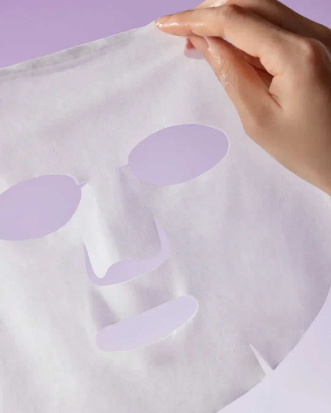 Hand holding a white facial sheet mask with cut-outs for eyes, nose, and mouth on a lavender background