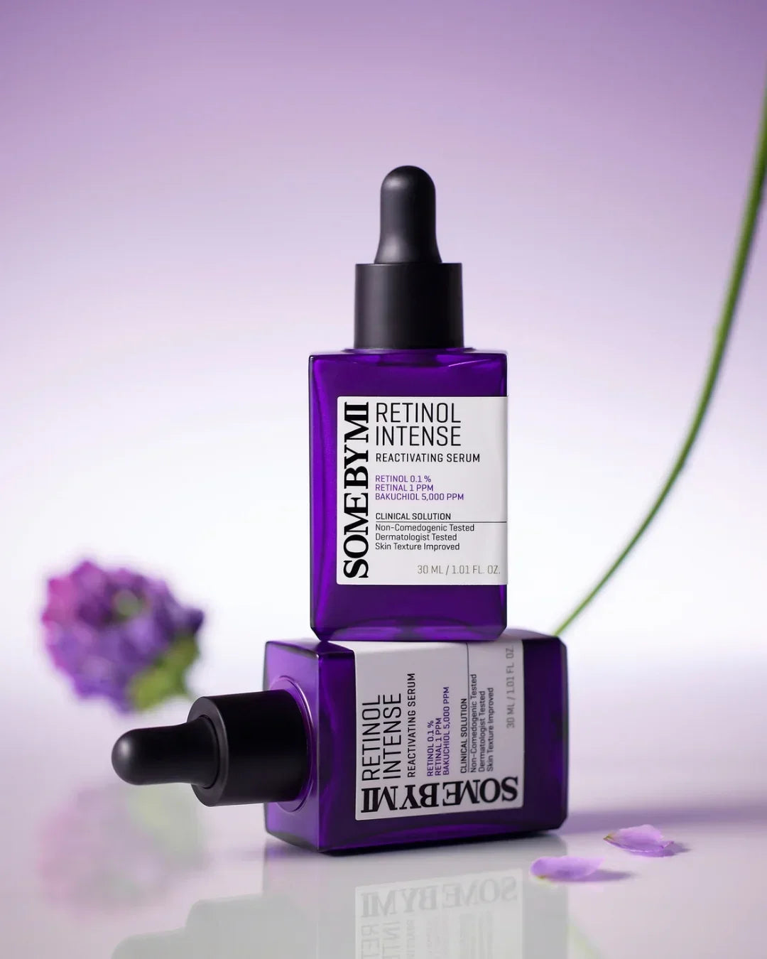Two purple bottles of Some By Mi Retinol Intense Reactivating Serum with black droppers on a reflective surface and a blurred purple flower