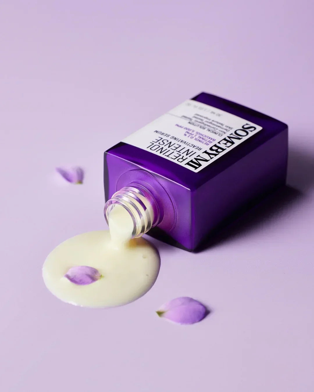 Purple bottle of Some By Mi Retinol Intense Reactivating Serum spilled on purple surface with cream and flower petals