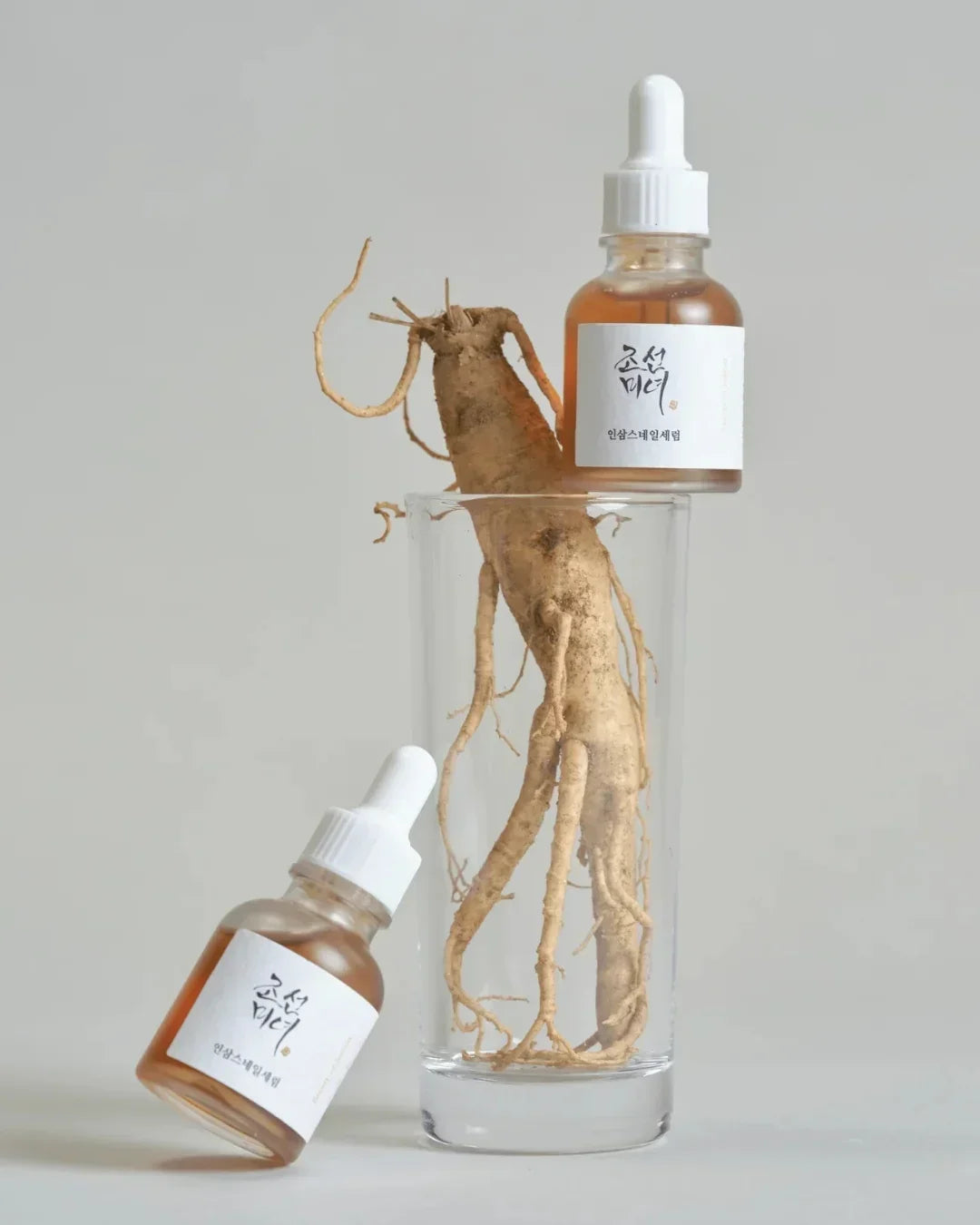 Glass with fresh ginseng root with two amber serum bottles featuring white dropper caps and labels