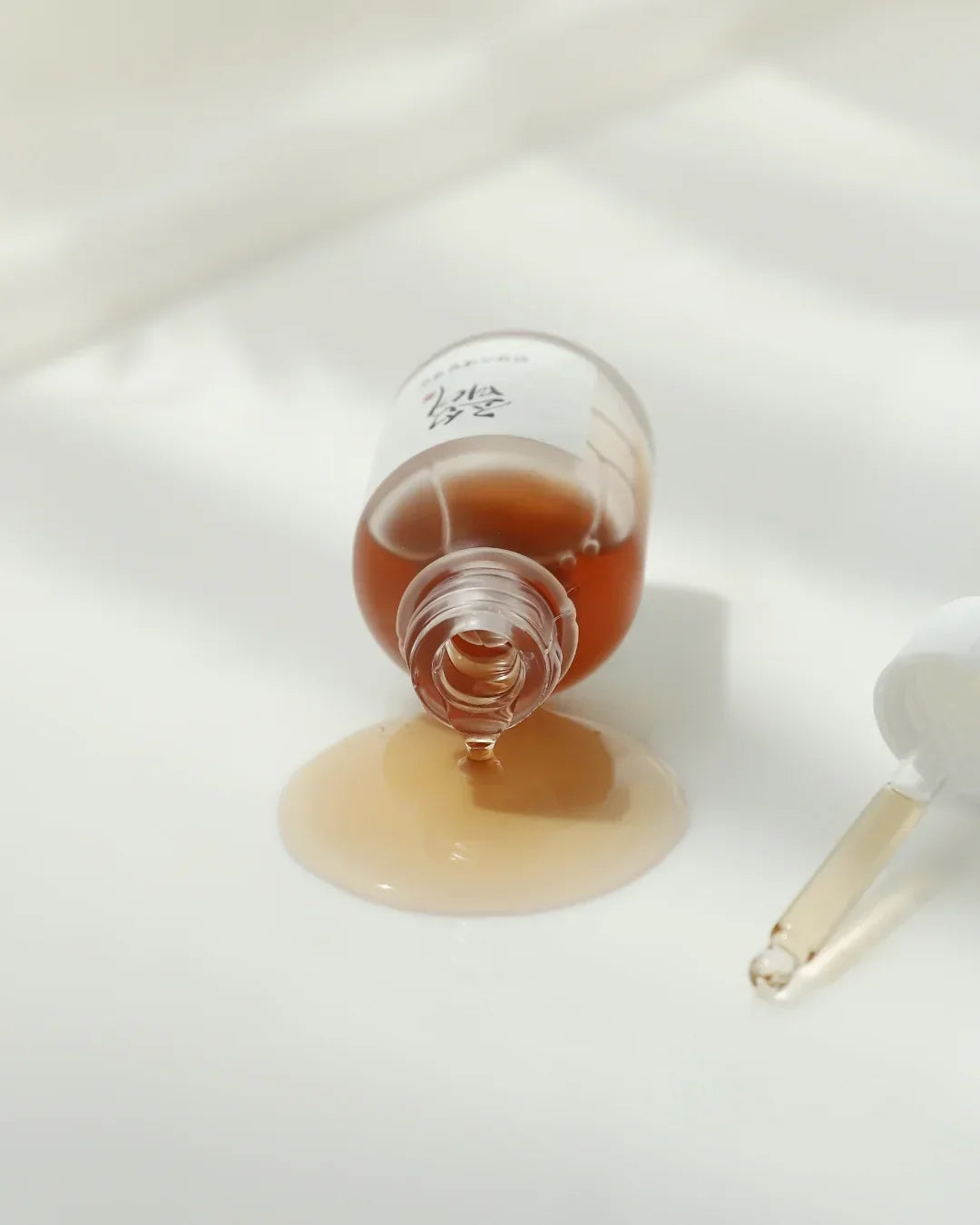 Brown glass serum bottle tipped over with spilled liquid and dropper on white surface