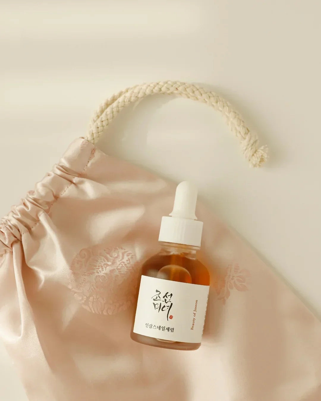 Glass dropper bottle of Korean ginseng snail mucin serum on blush pink satin pouch with braided drawstring