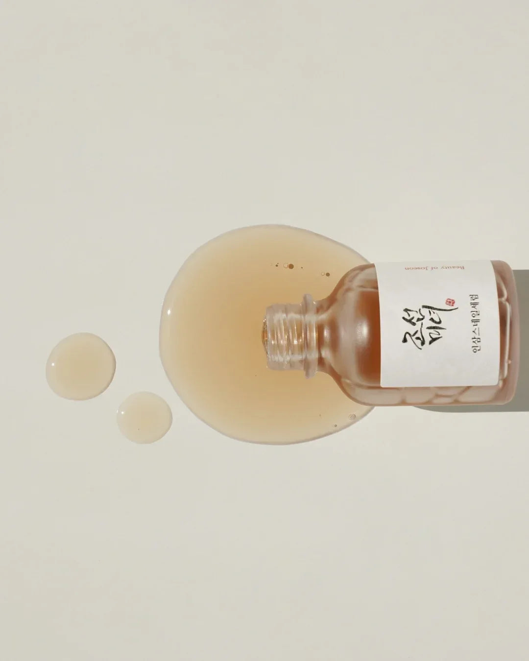 Open brown frosted glass bottle spilling light beige serum on white surface with Korean text label