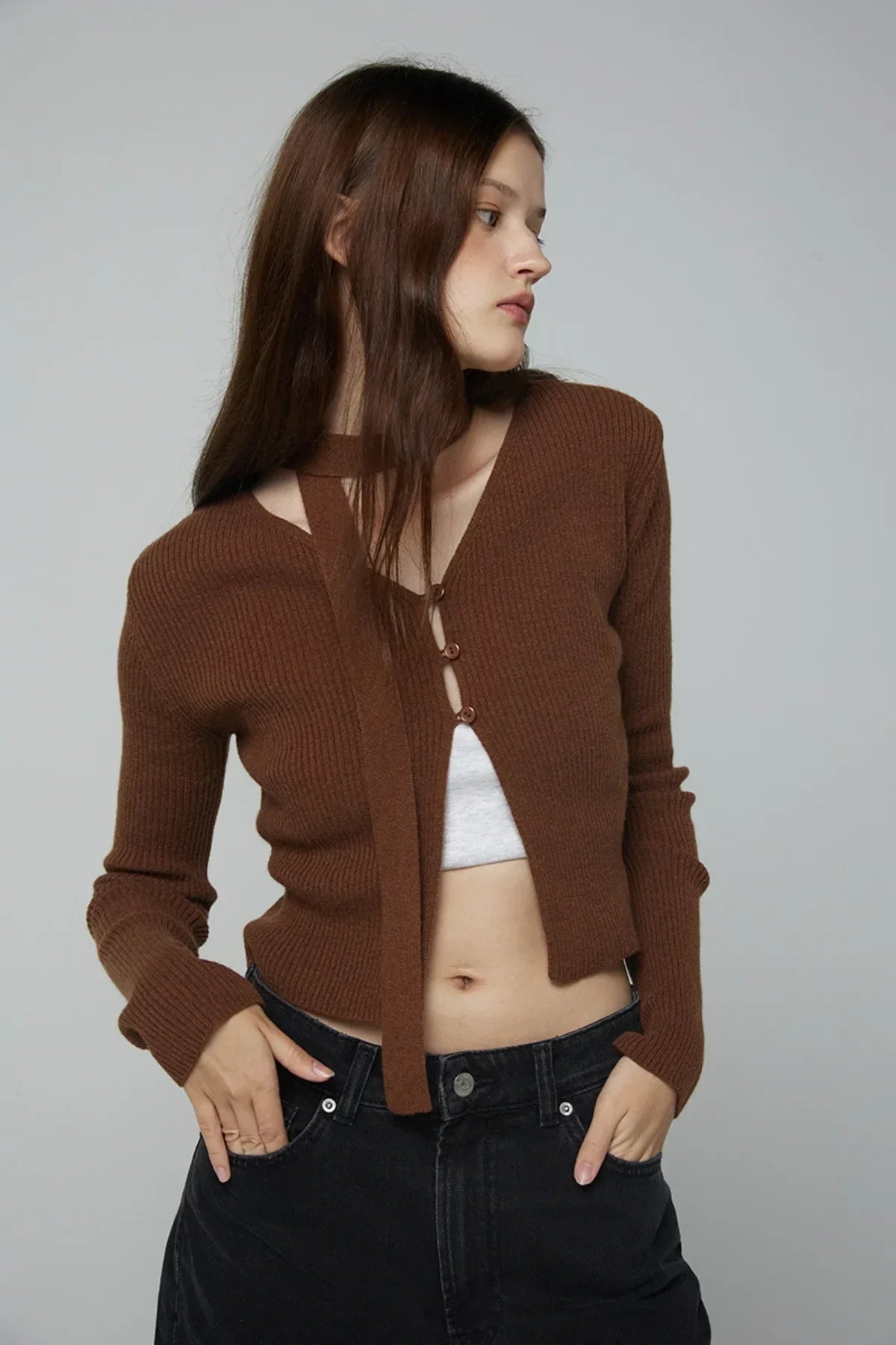 Woman wearing brown ribbed V-neck open cardigan with buttons and black jeans, posing against gray background