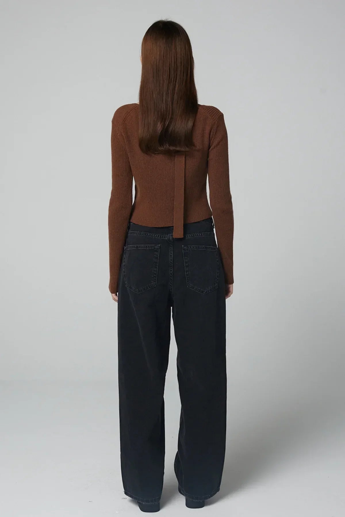 Back view of woman wearing ribbed brown V-neck cardigan and black wide-leg jeans on gray background