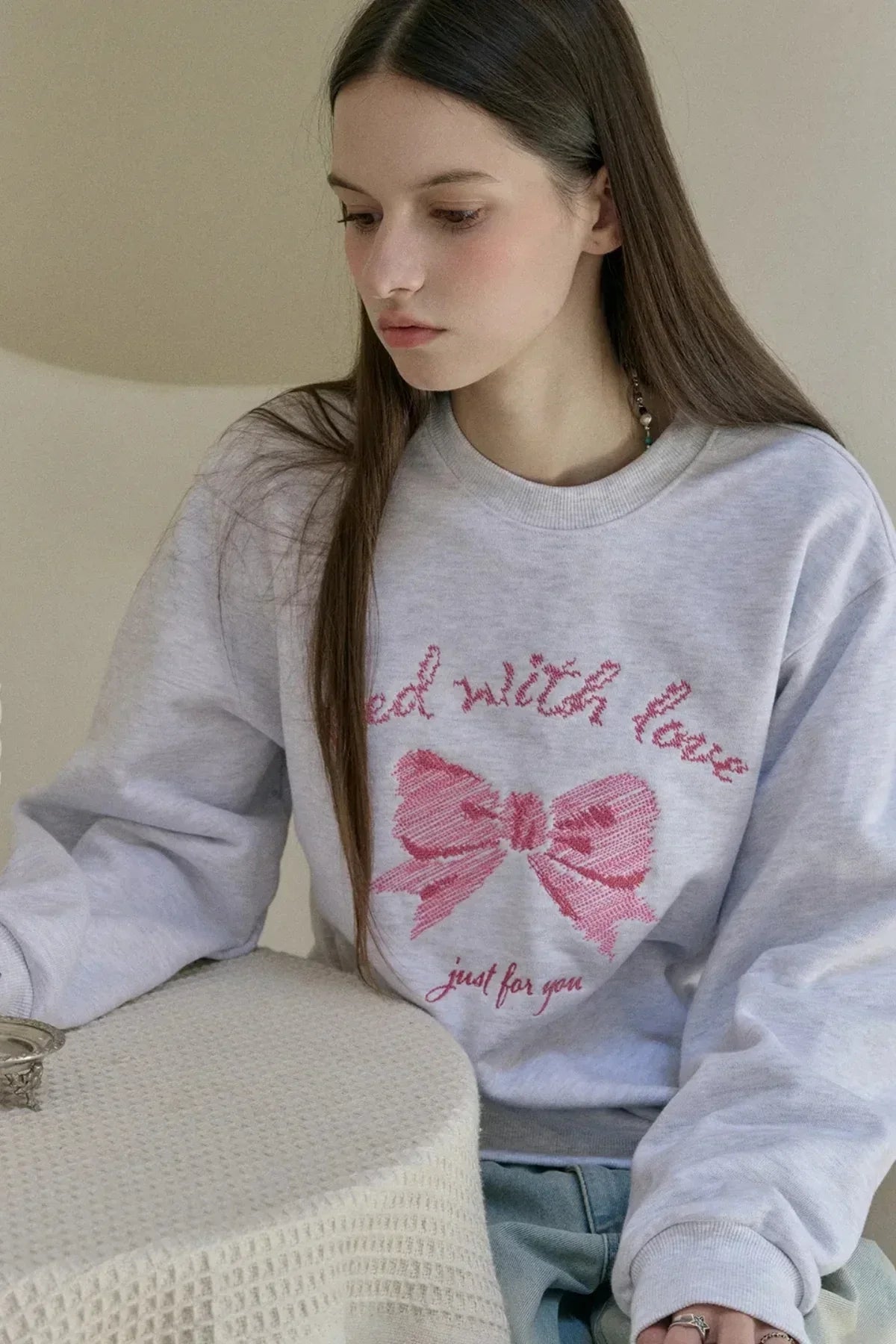 Young woman wearing a melange white sweatshirt with pink embroidered bow and text, sitting indoors