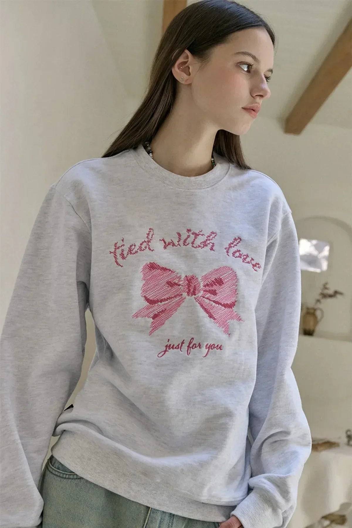 Woman wearing light grey sweatshirt with pink embroidered bow and text tied with love just for you