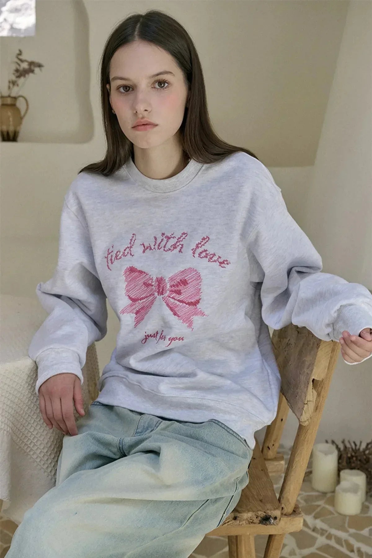 Young woman wearing light grey sweatshirt with pink bow embroidery and text sitting on wooden chair