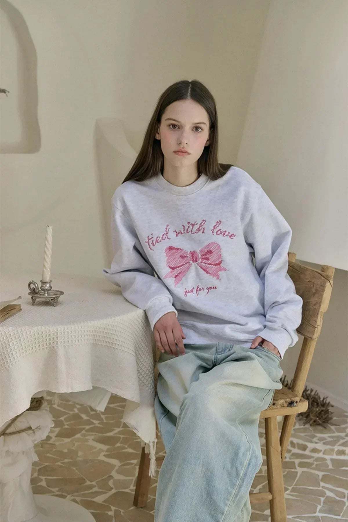 Young woman wearing light gray sweatshirt with pink bow embroidery and text, seated on wooden chair indoors