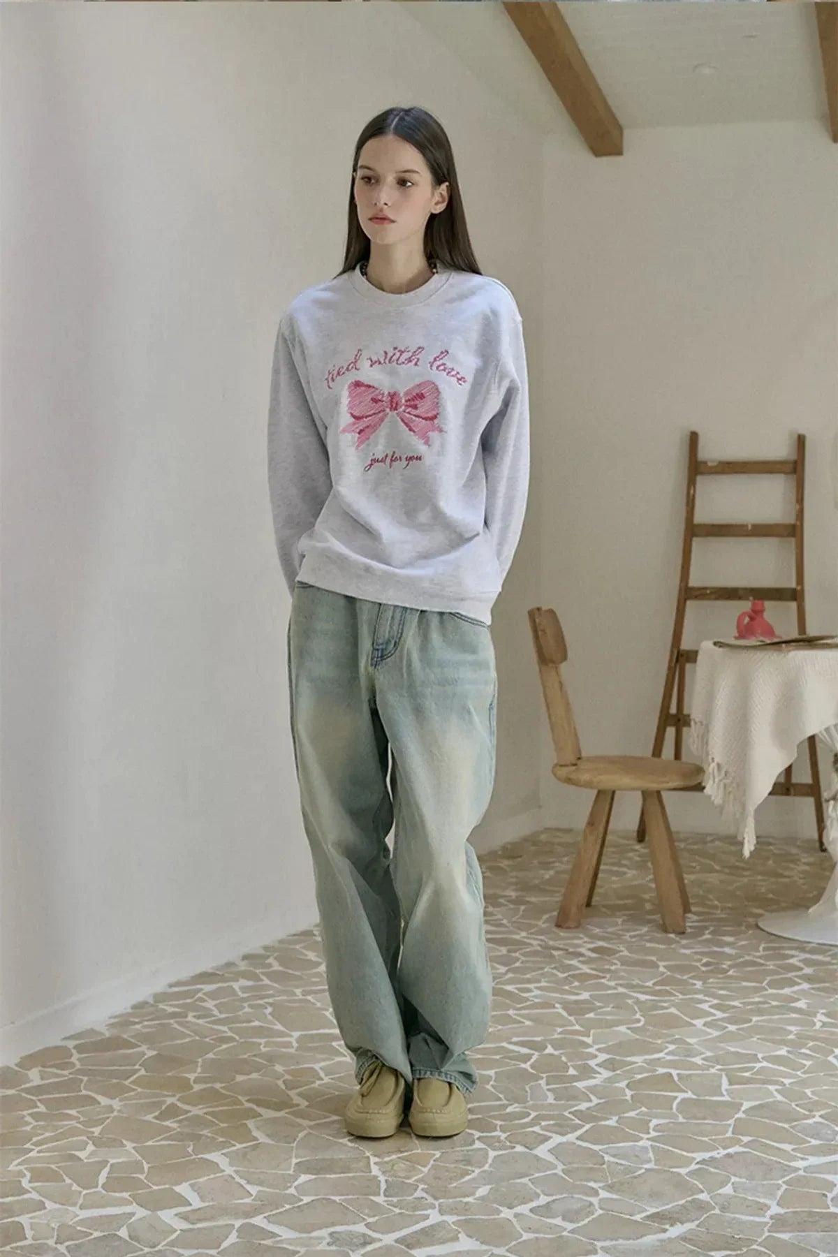 Young woman wearing a melange white sweatshirt with pink bow embroidery and loose jeans in a minimalist room