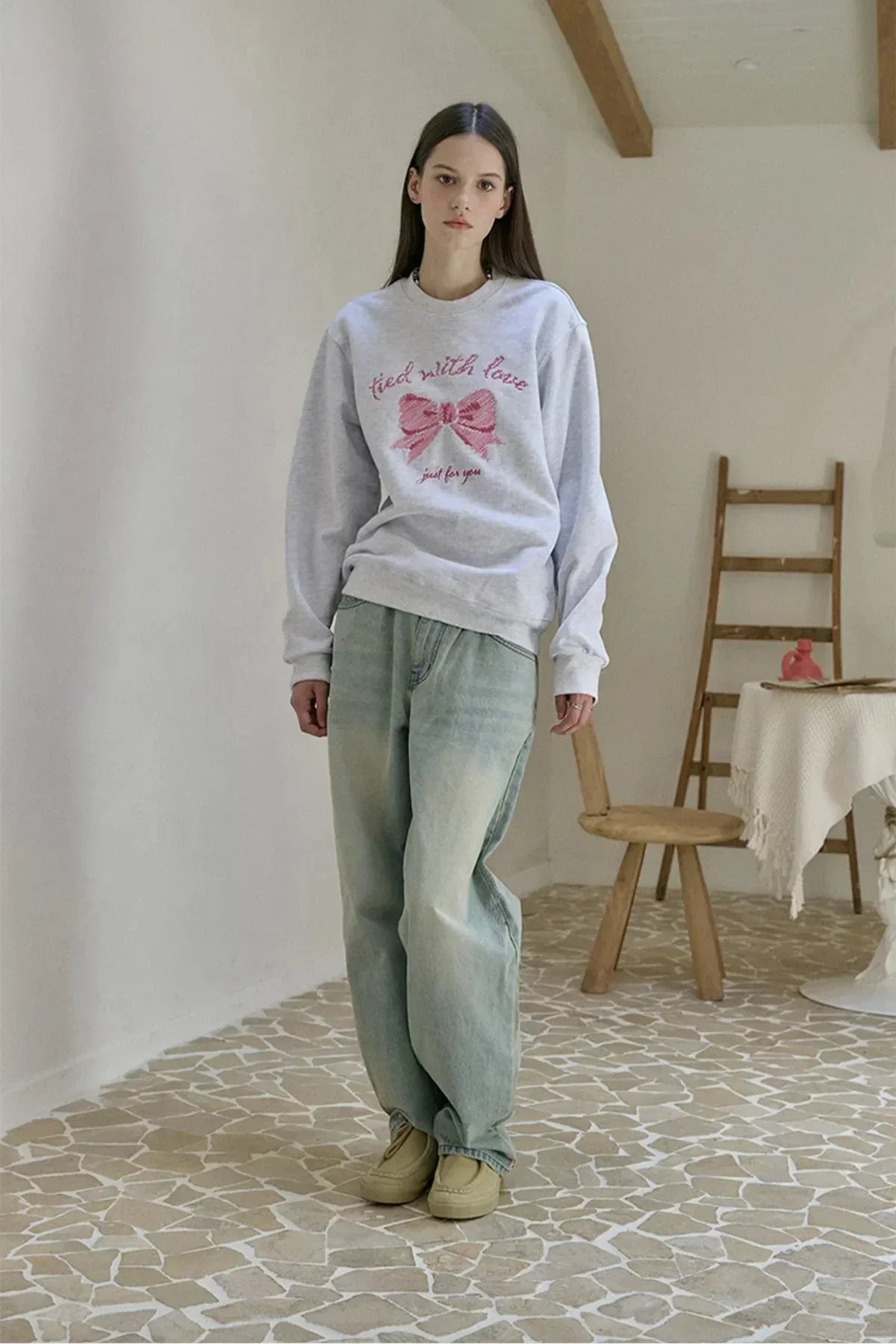 Young woman wearing white sweatshirt with pink bow embroidery and light blue jeans in a minimalist room