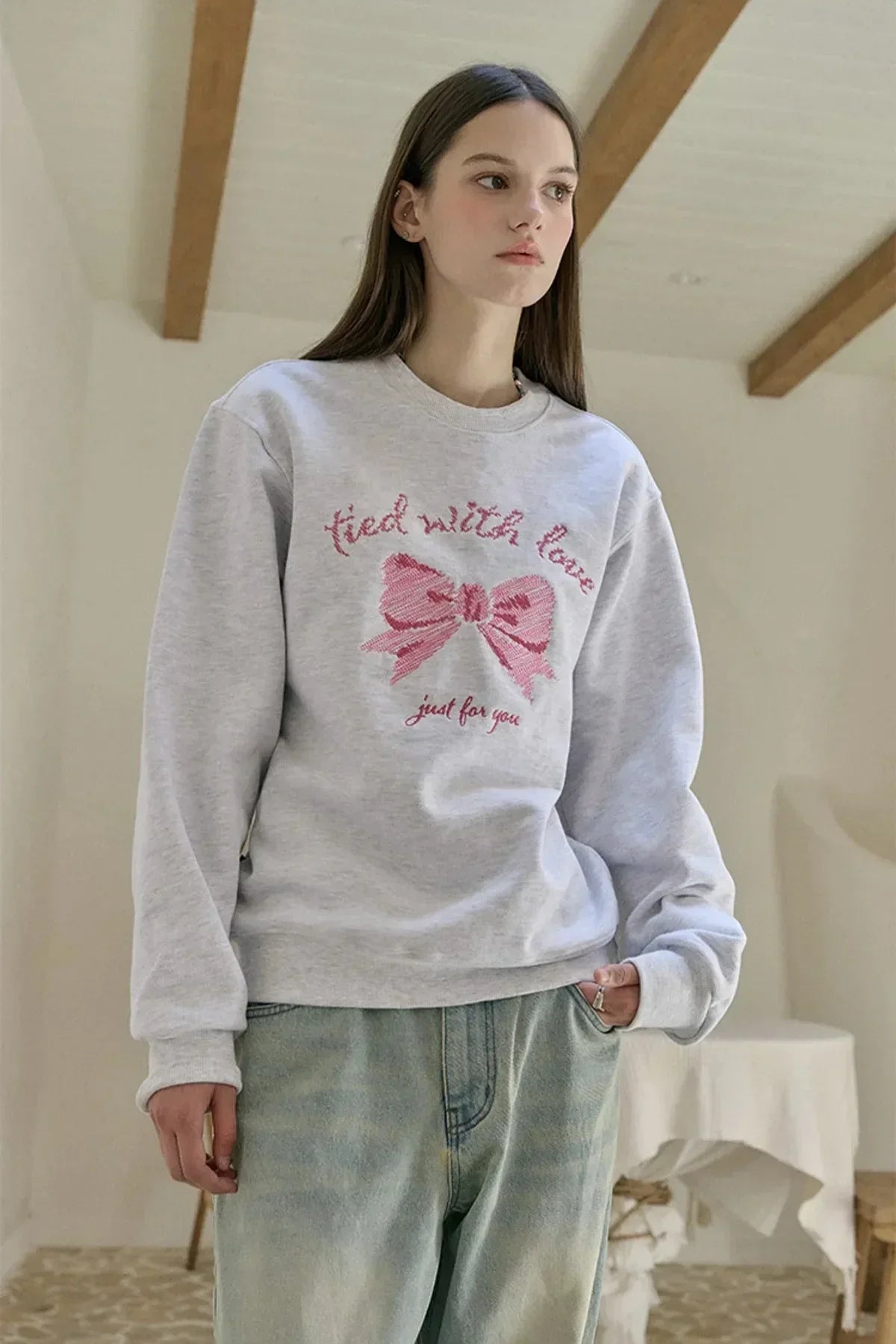 Young woman wearing light gray sweatshirt with pink embroidered bow and text tied with love
