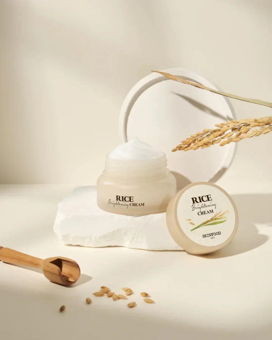 Open jar of Skinfood Rice Brightening Cream with cream texture, rice grains, wooden scoop, and rice stalk on white background