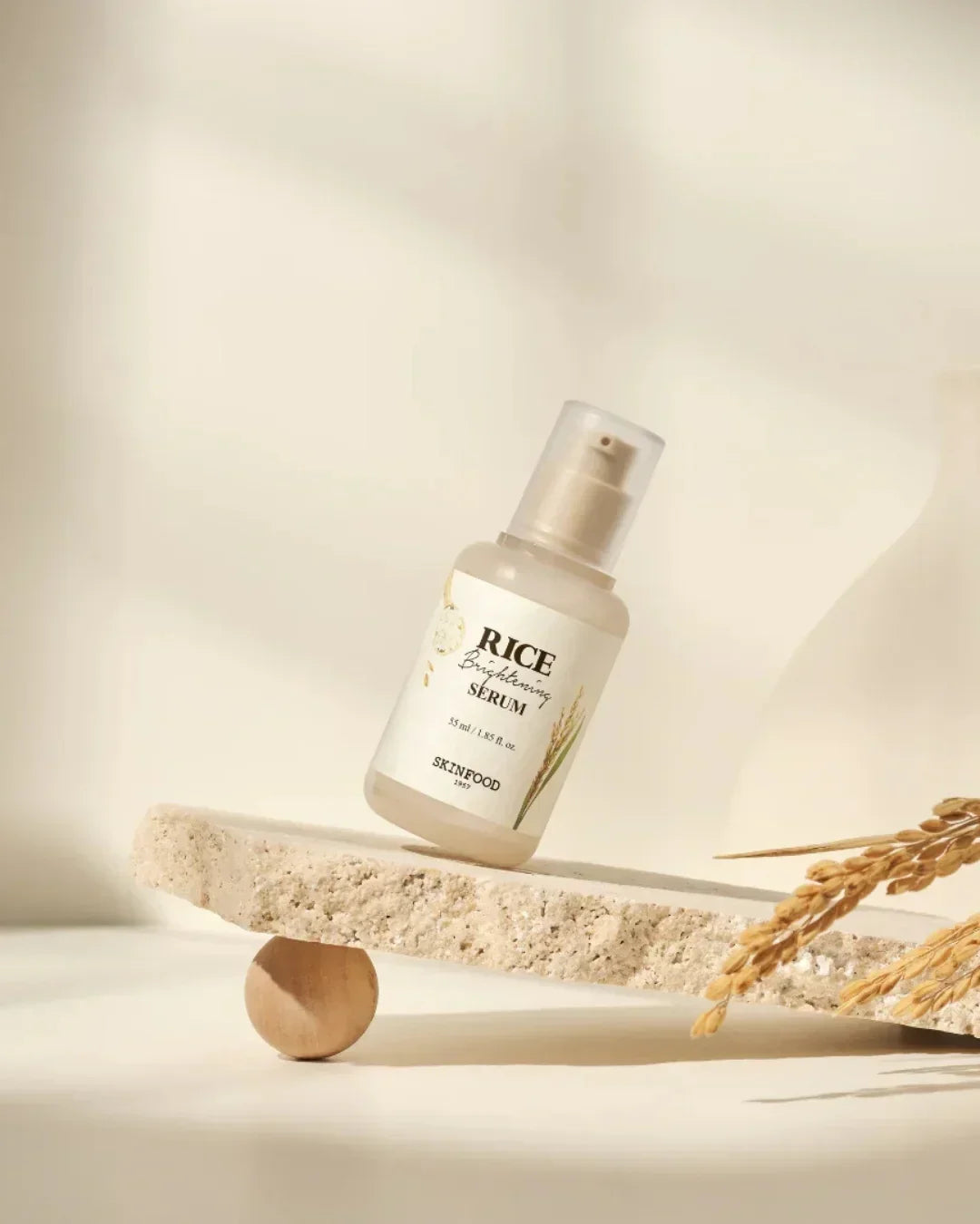 Skinfood Rice Brightening Serum 55ml bottle on textured stone with rice stalks, studio light