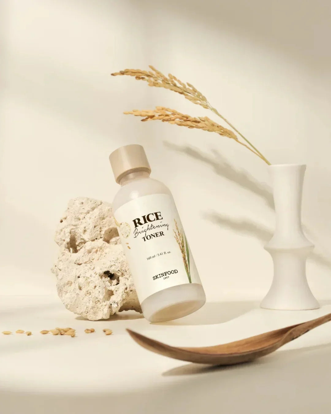 Bottle of SkinFood Rice Brightening Toner with natural rice stalks, stone, and wooden spoon on beige background