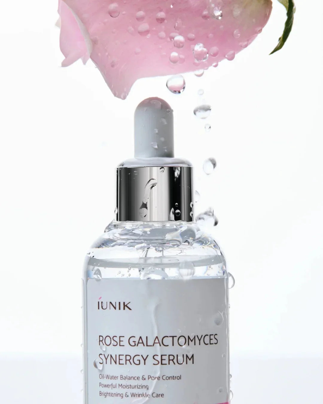 iUNIK Rose Galactomyces Synergy Serum bottle with dropper under pink rose petals and water droplets