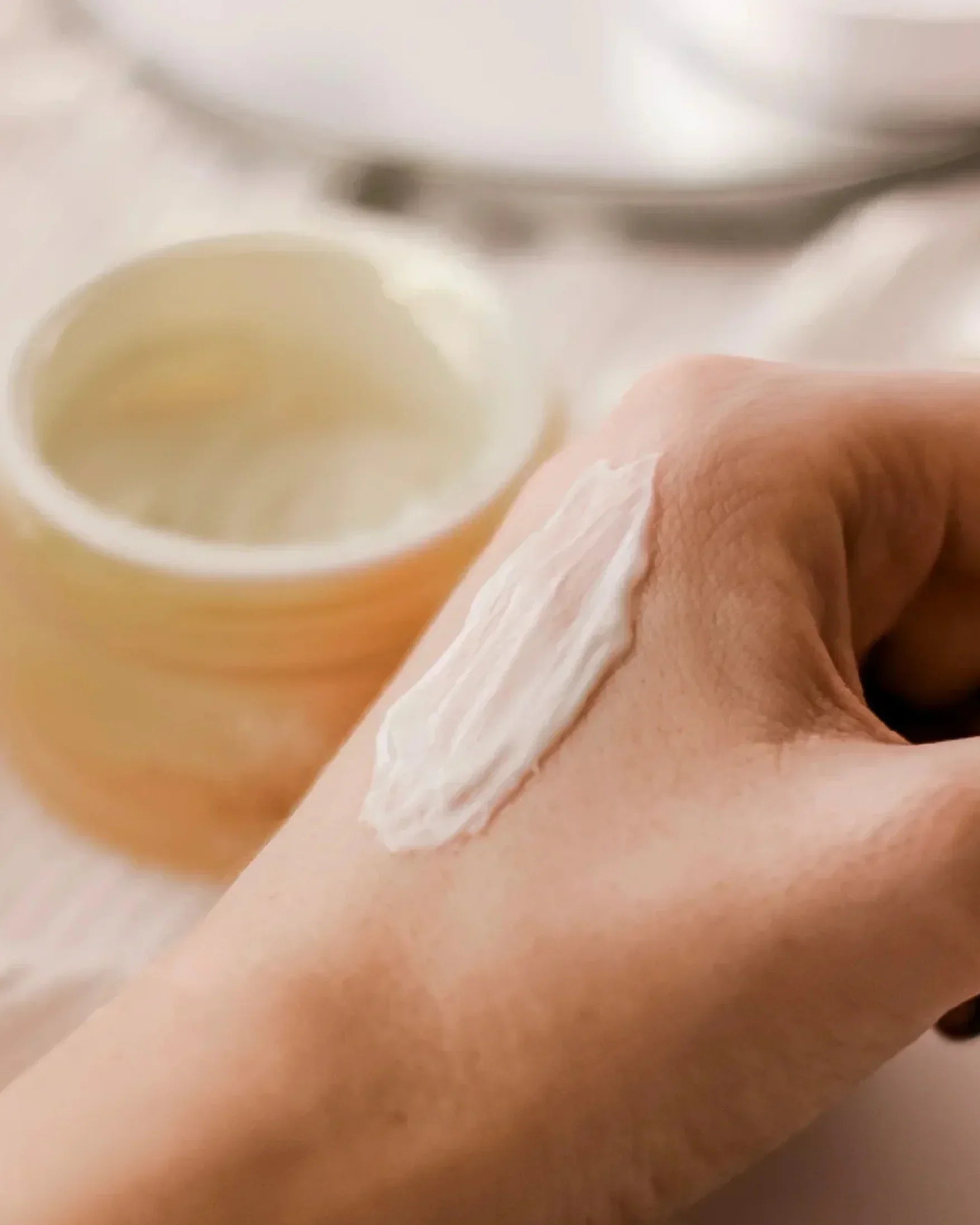 Hand with white propolis cream applied, blurred cream jar in background on light surface