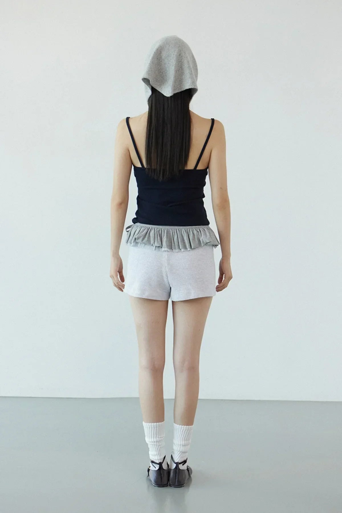 Woman wearing navy ruffle bra top, grey shorts, white socks, black shoes, and grey headscarf indoors