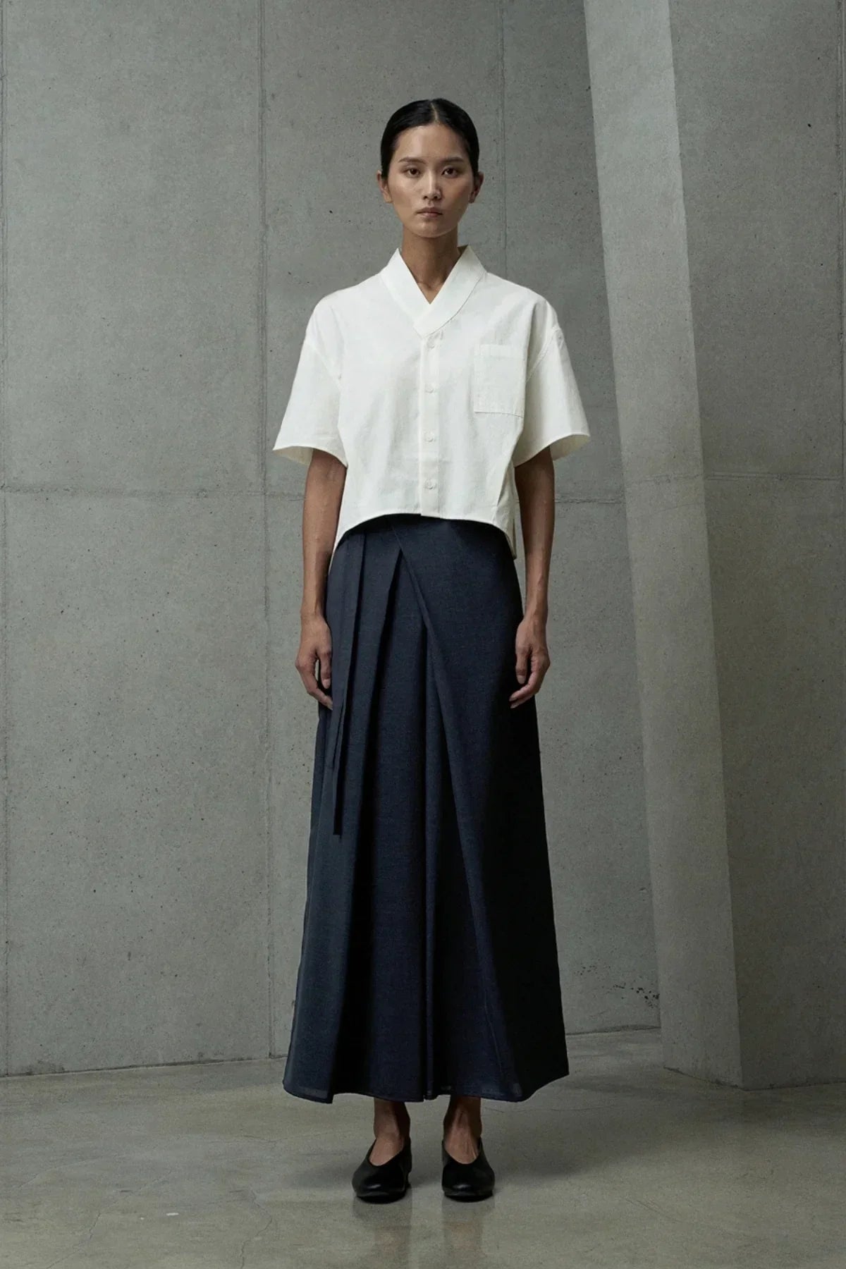 Model wearing white short-sleeve button-up shirt and navy blue pleated wrap skirt in minimalist concrete setting