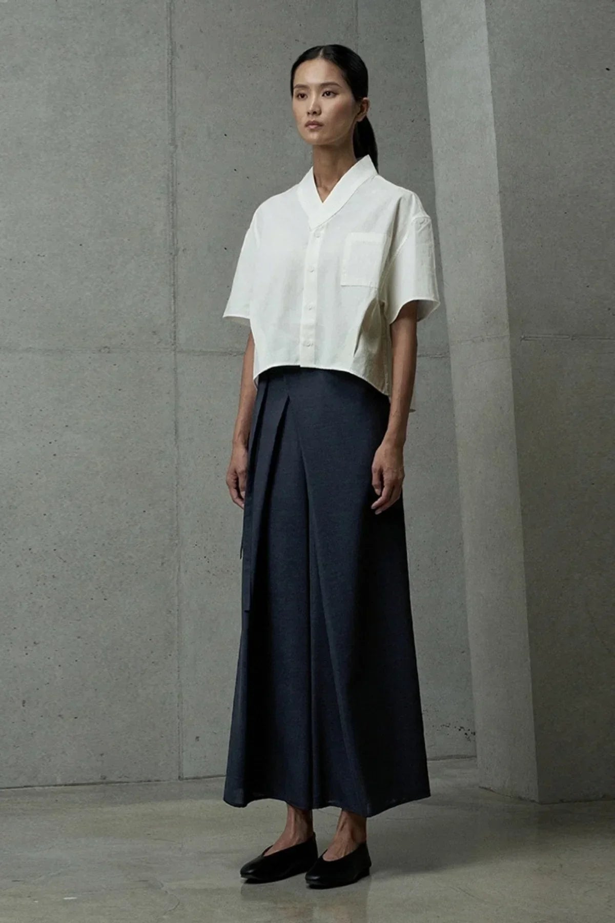 Model wearing cropped white button-up shirt and navy blue wide-leg pants in minimalist concrete setting
