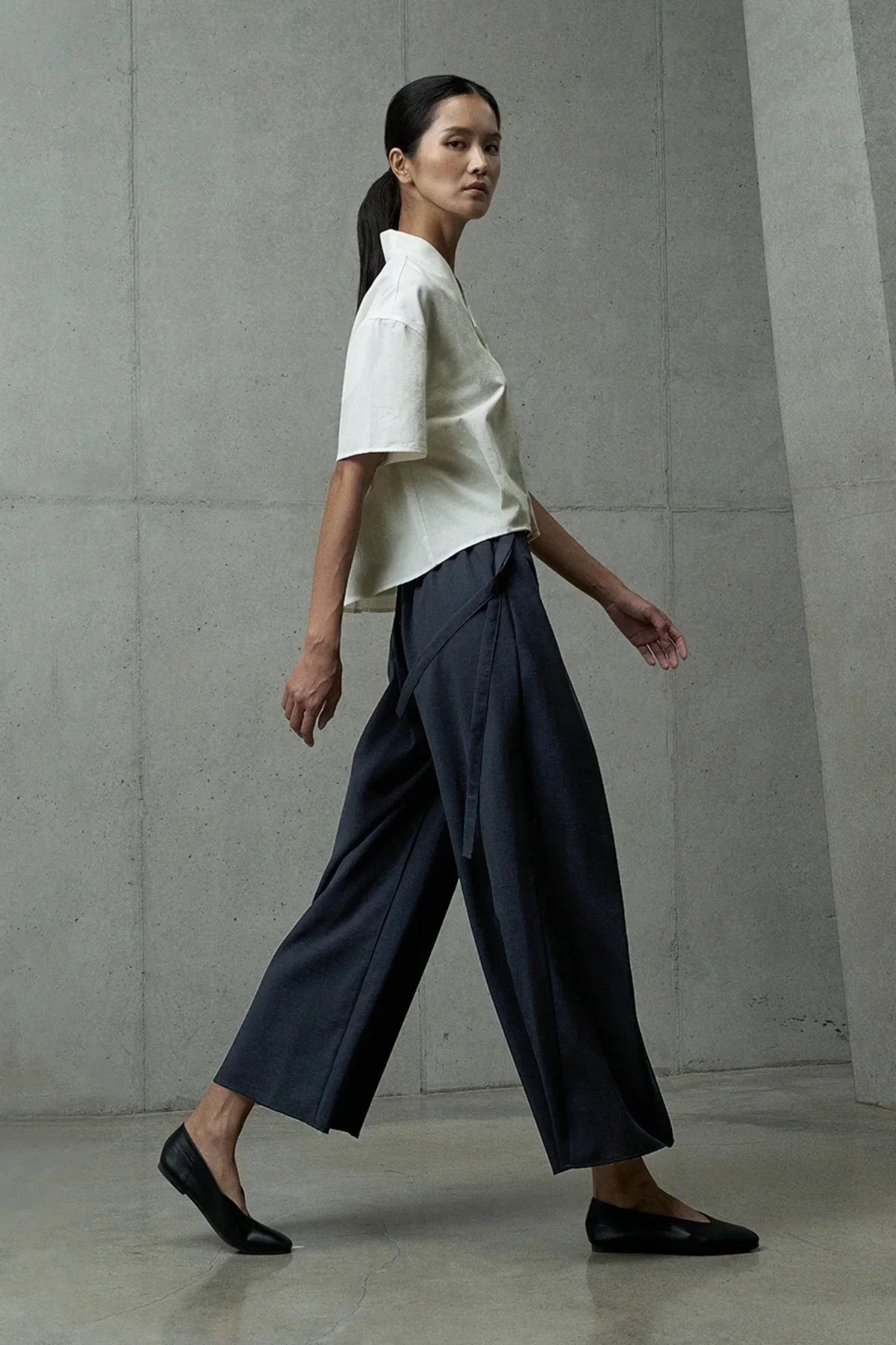 Woman walking in studio wearing navy blue wide-leg pants, white short-sleeve top, and black flats