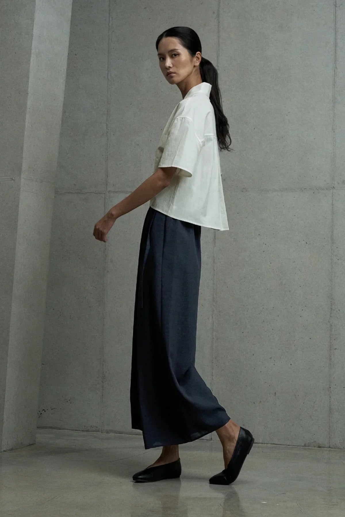 Woman wearing navy blue wide-leg pants and white short-sleeve shirt in minimal concrete studio