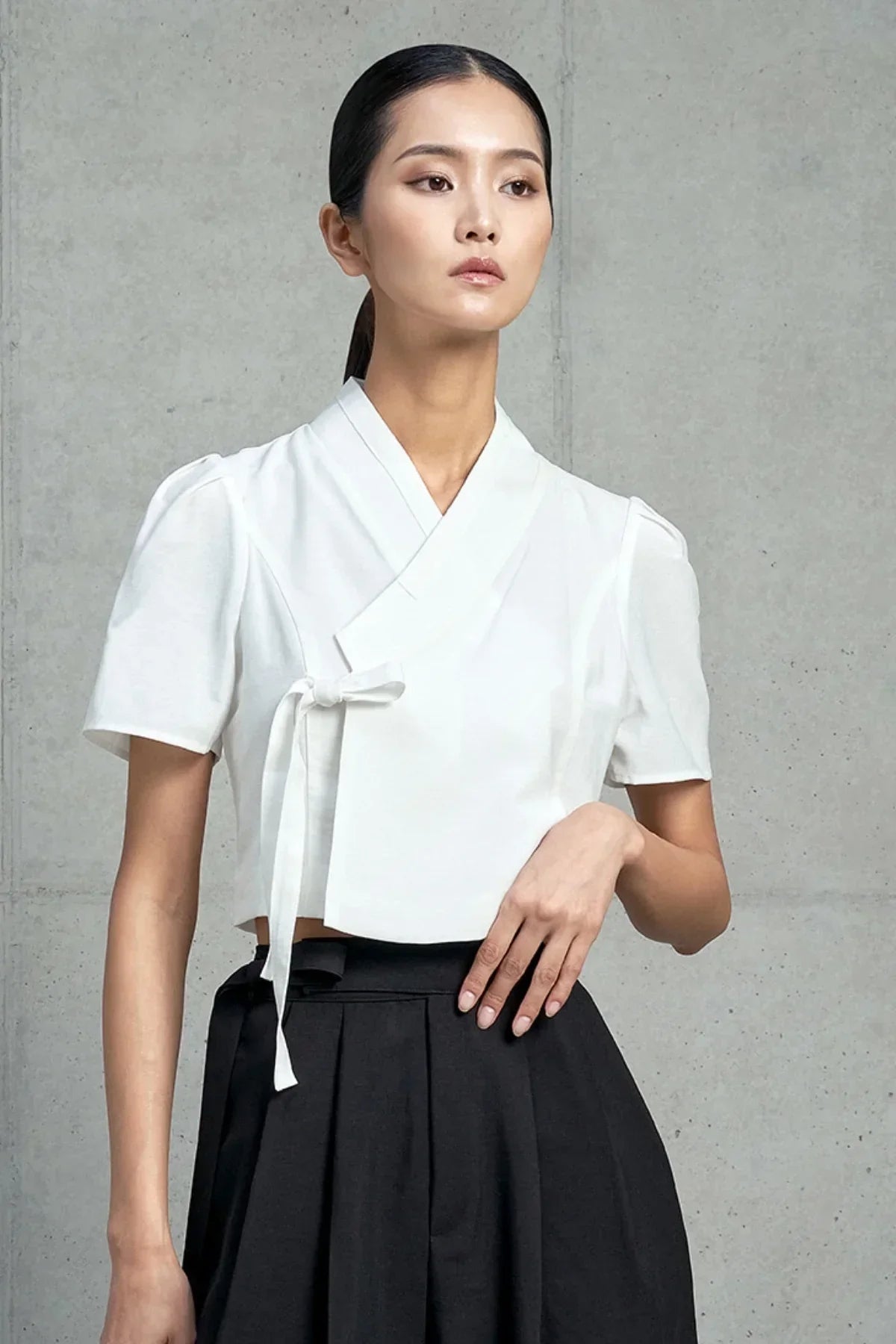 Woman wearing a white short-sleeved cropped jeogori with a tie detail and black pleated skirt