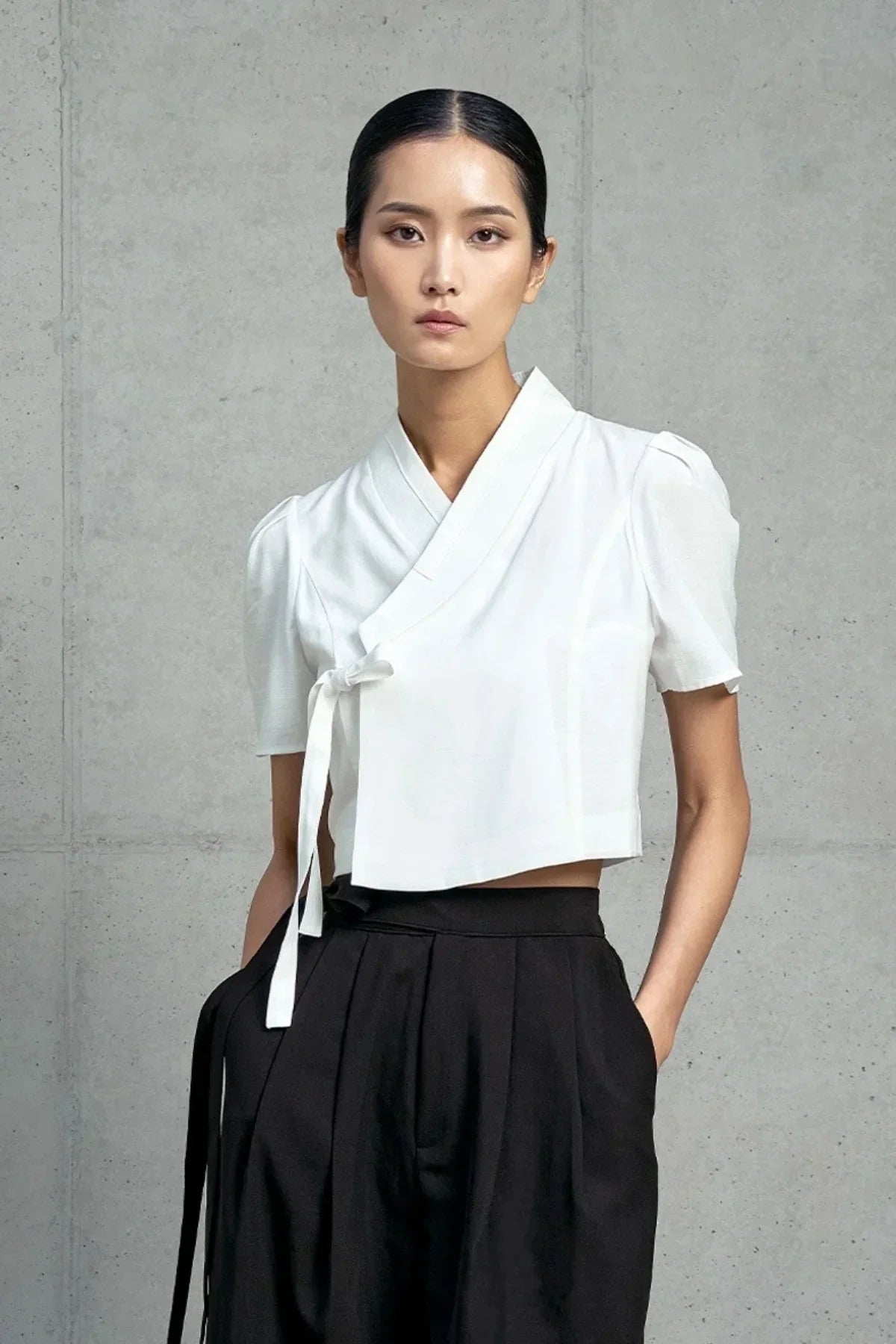 Asian woman wearing white short-sleeved wrap top and high-waisted black pants against gray wall