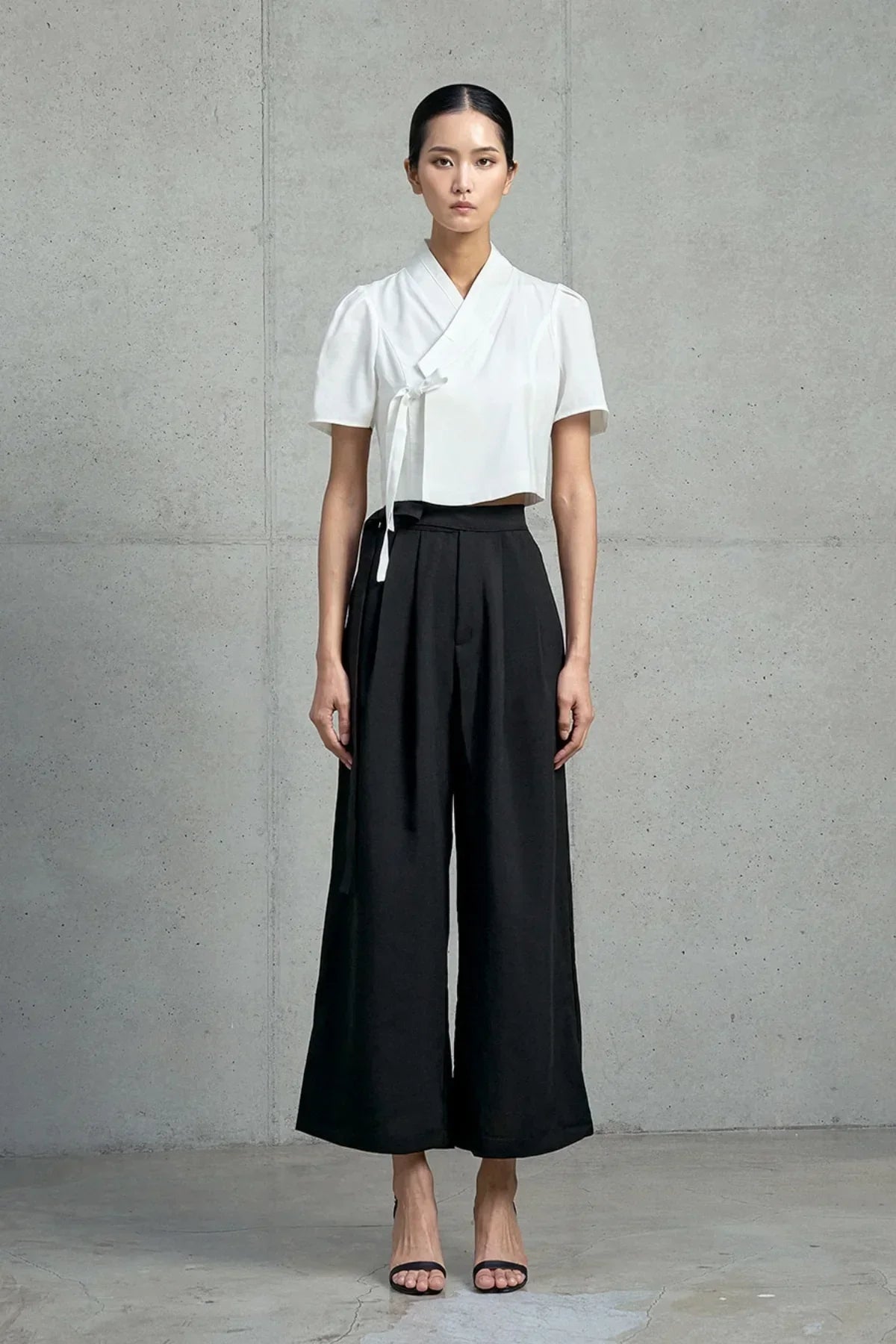 Woman wearing a white short-sleeved crop jeogori and black wide-leg pants against a gray concrete wall