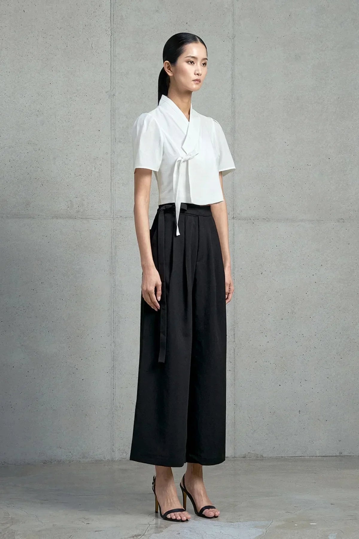 Model wearing a white short-sleeved jeogori and high-waisted black wide-leg pants against a concrete wall