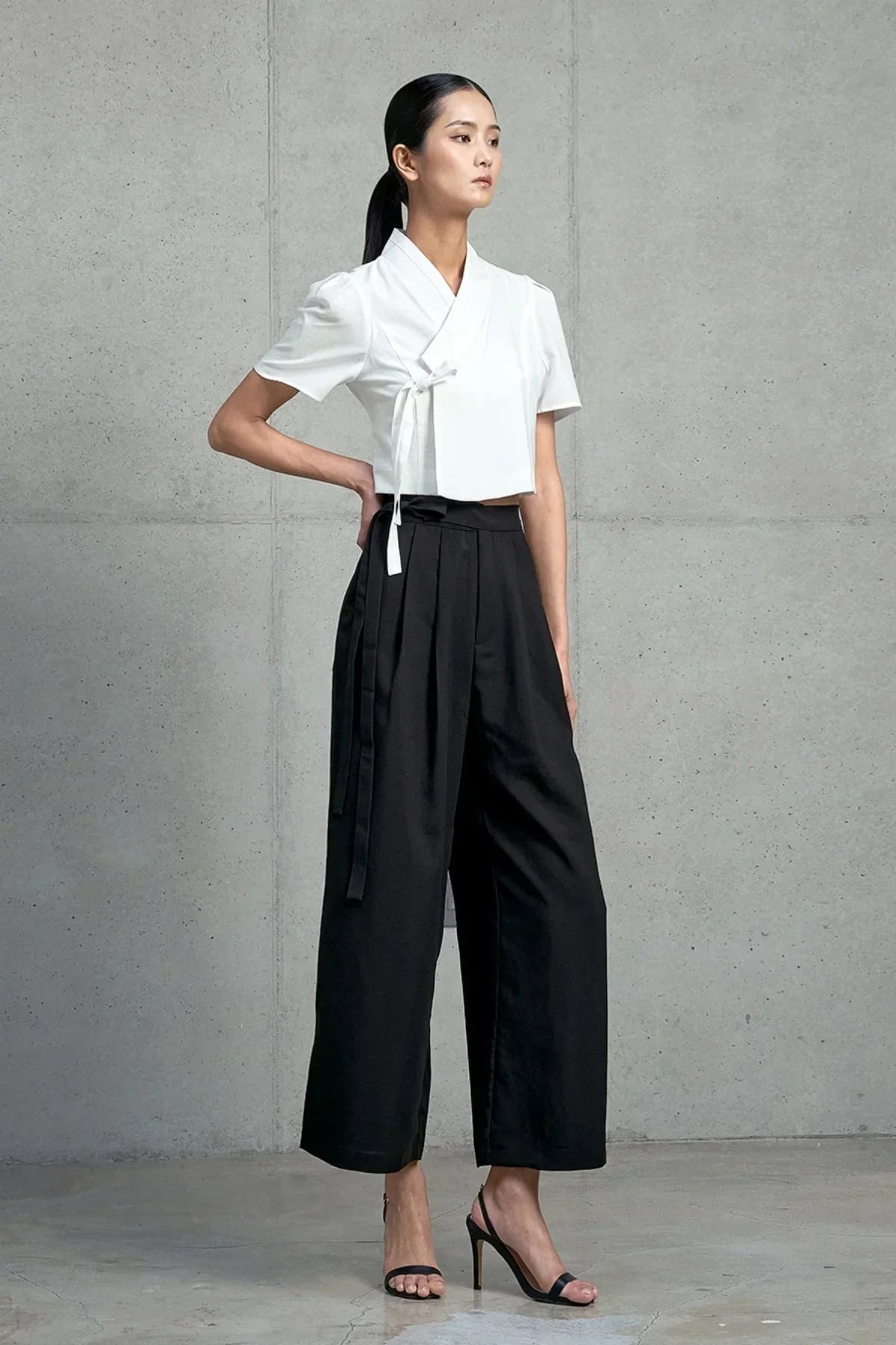 Model wearing a white short-sleeved jeogori top and black wide-leg pants with high heels against a gray concrete wall