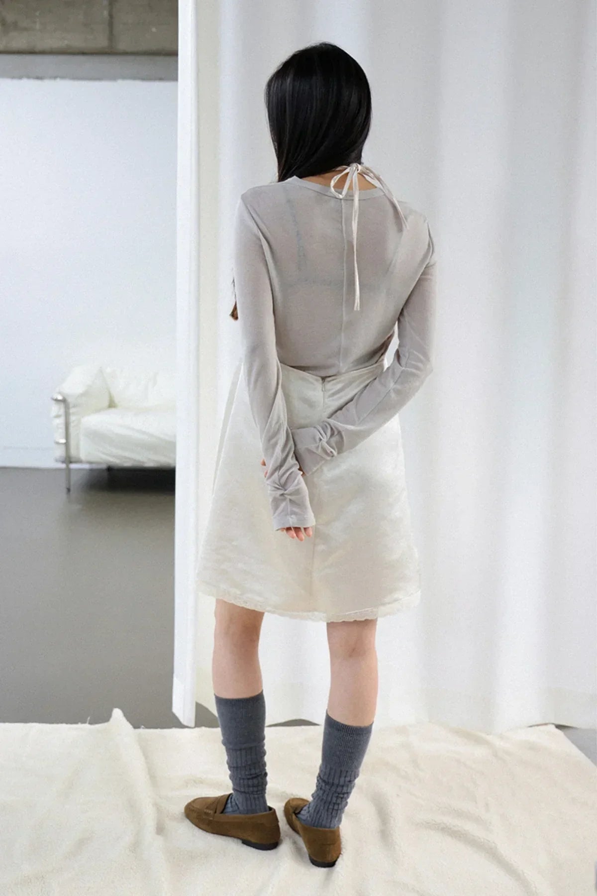 Woman wearing ivory satin mini skirt and gray long-sleeve top with tie back, standing indoors