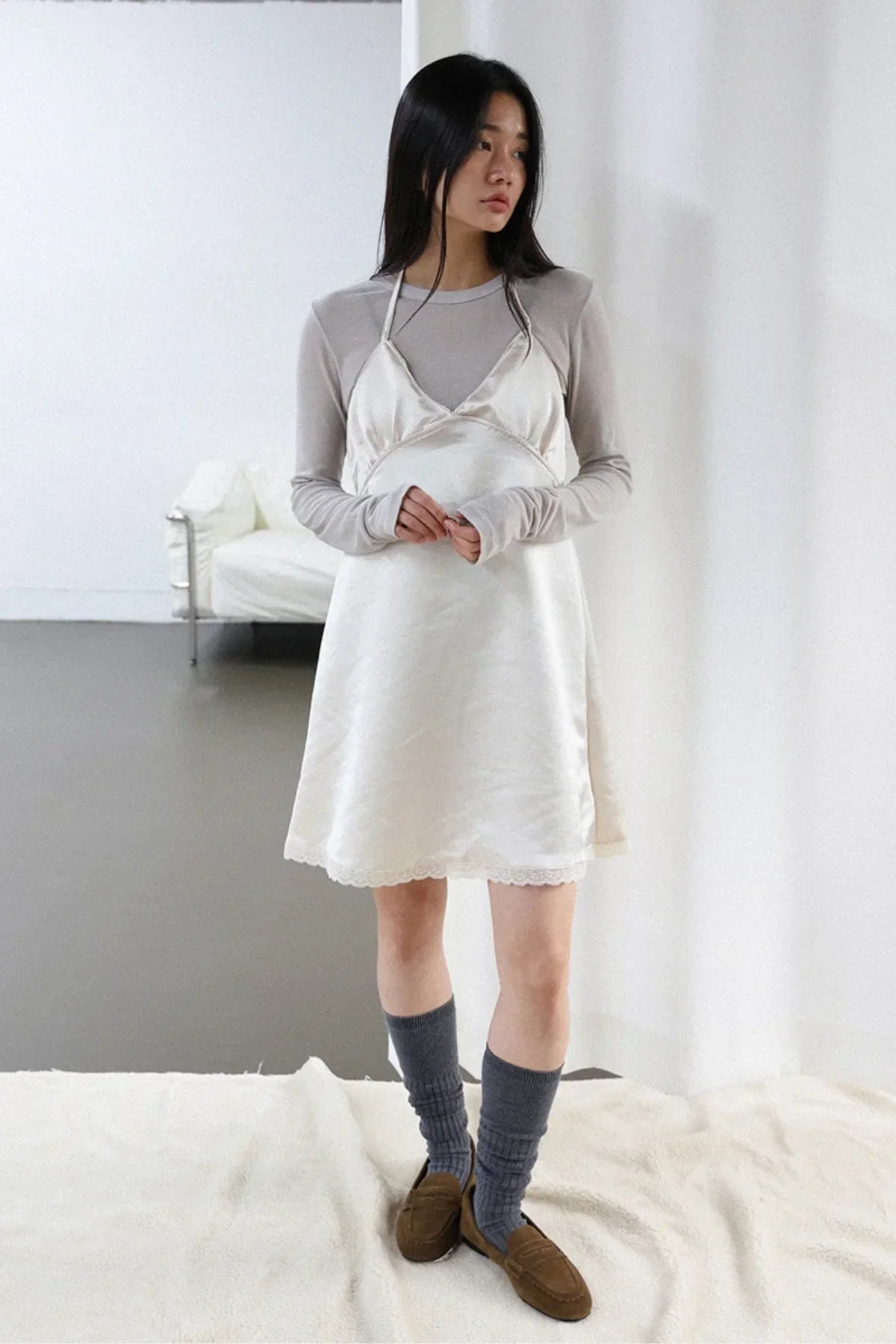 Young woman wearing ivory satin mini dress layered over a long sleeve shirt with gray socks and brown loafers indoors