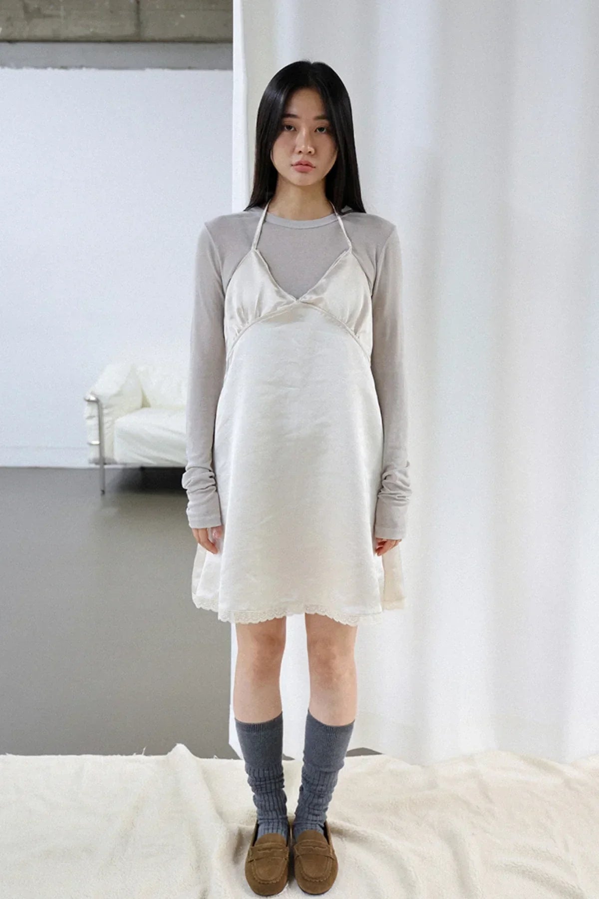 Woman wearing ivory satin mini dress layered over gray long sleeve top, paired with gray socks and brown loafers in minimalist room