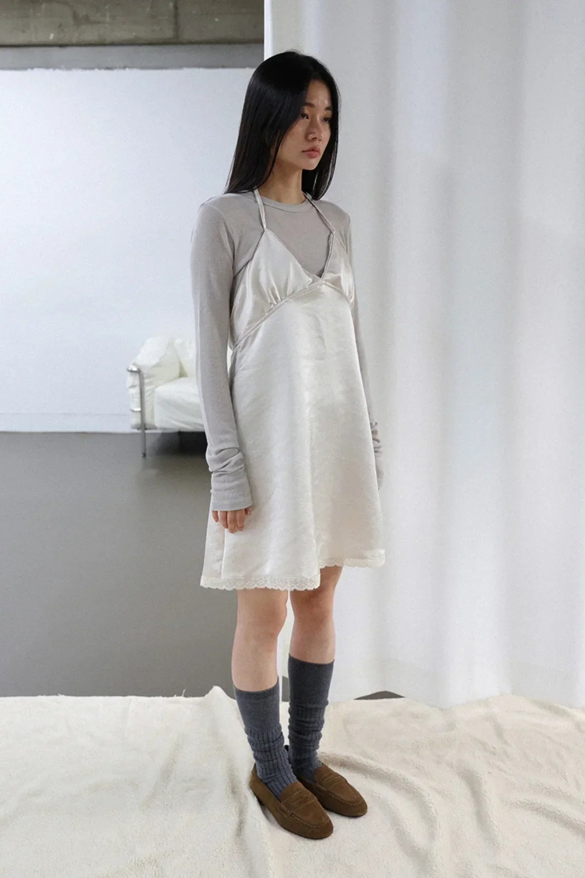 Model wearing ivory satin mini dress layered over gray long sleeve top with gray socks and brown shoes in minimalist room