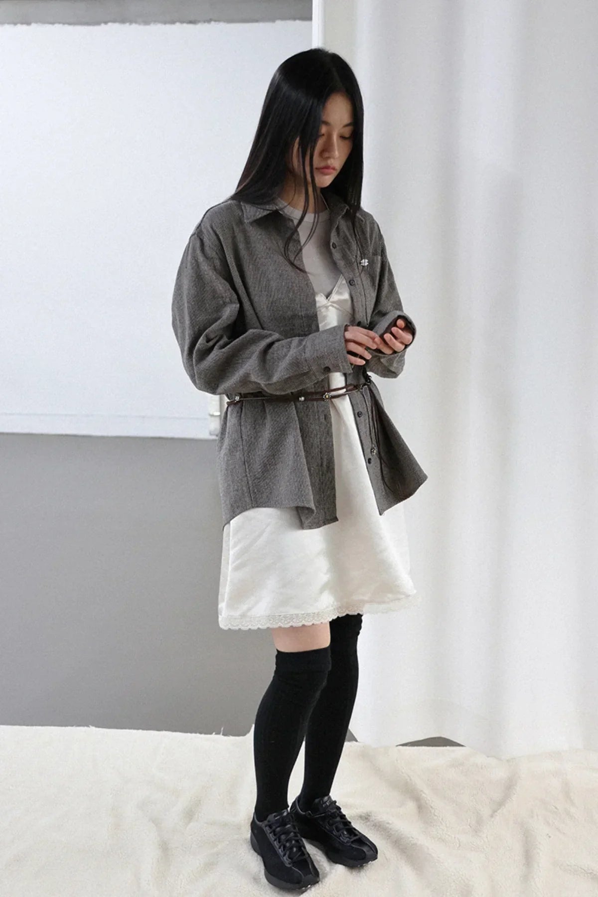 woman wearing ivory satin mini dress with gray overshirt, black knee-high socks, and black sneakers in studio