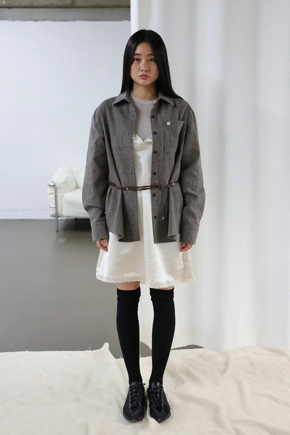 Woman wearing ivory satin mini dress with gray shirt layered, black thigh-high socks, and black shoes in minimalist studio