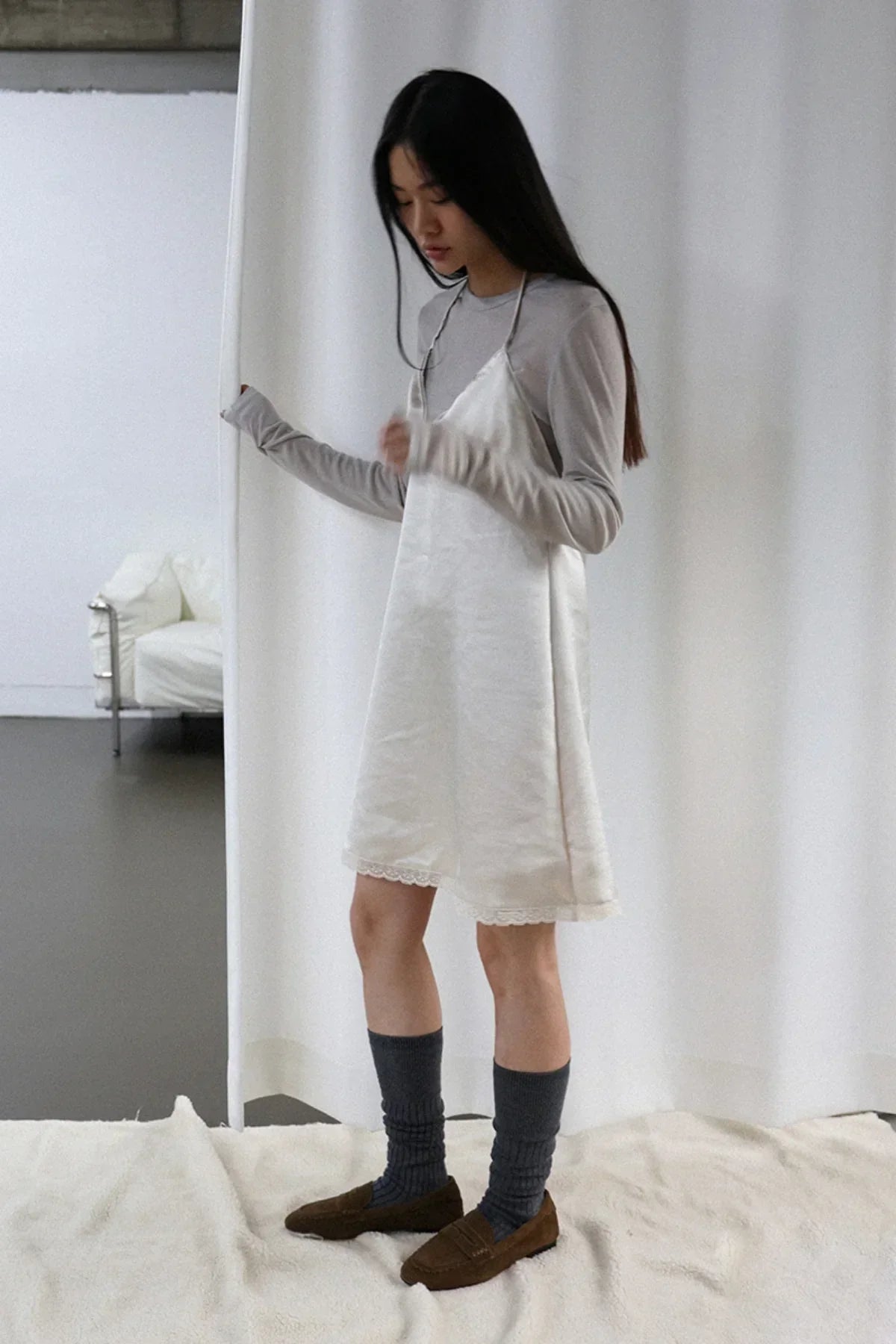 Woman wearing ivory satin mini dress over gray long-sleeve shirt with gray socks and brown loafers in minimalist setting