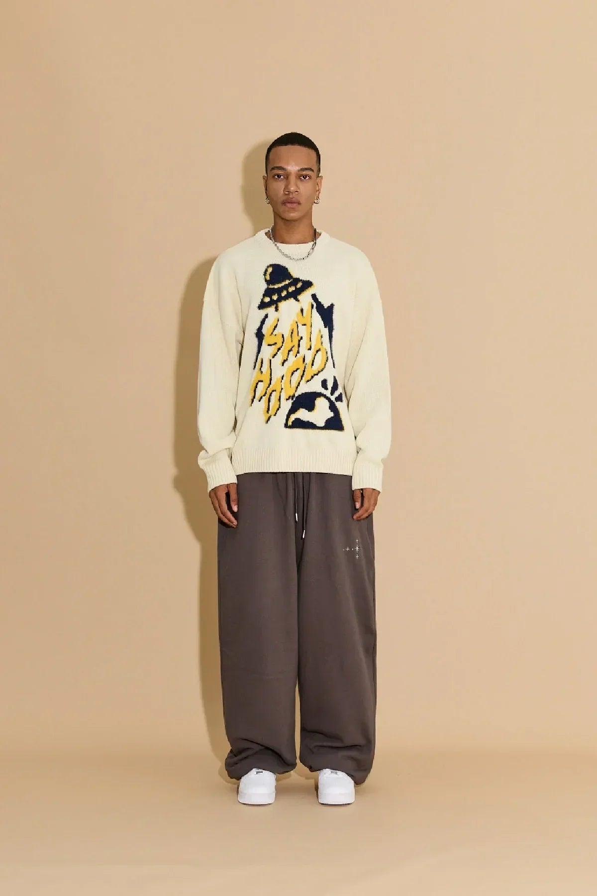 Model wearing ivory knit sweater with abstract graphic, loose brown pants, and white sneakers on tan background