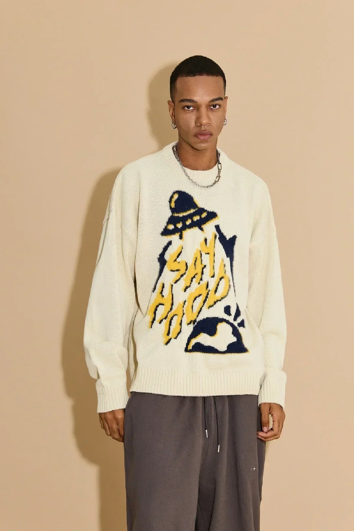 Male model wearing ivory knit sweater with yellow and black UFO graphic and text, paired with gray pants