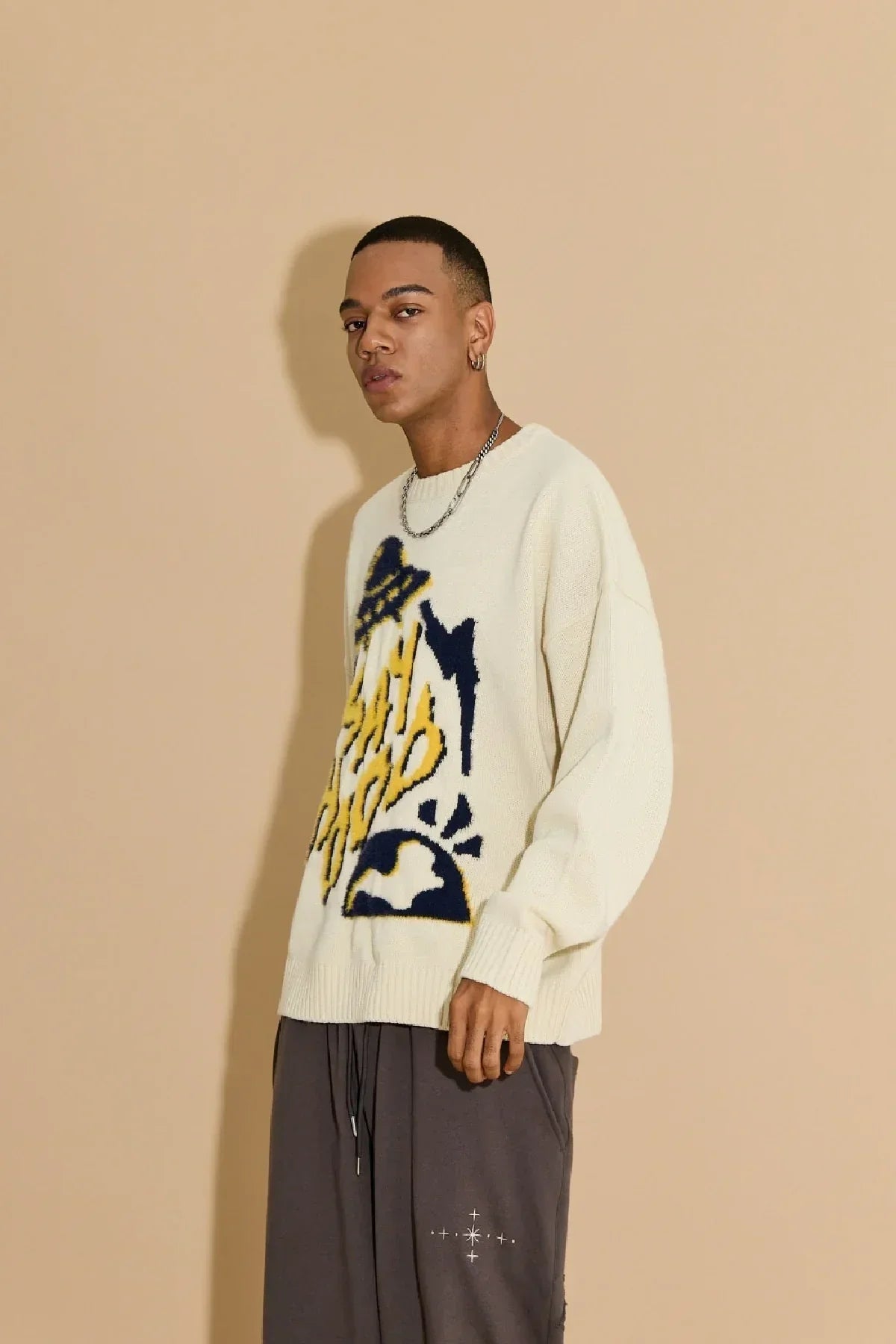 Fashion model wearing ivory knit sweater with yellow and navy graphic design, paired with dark gray pants