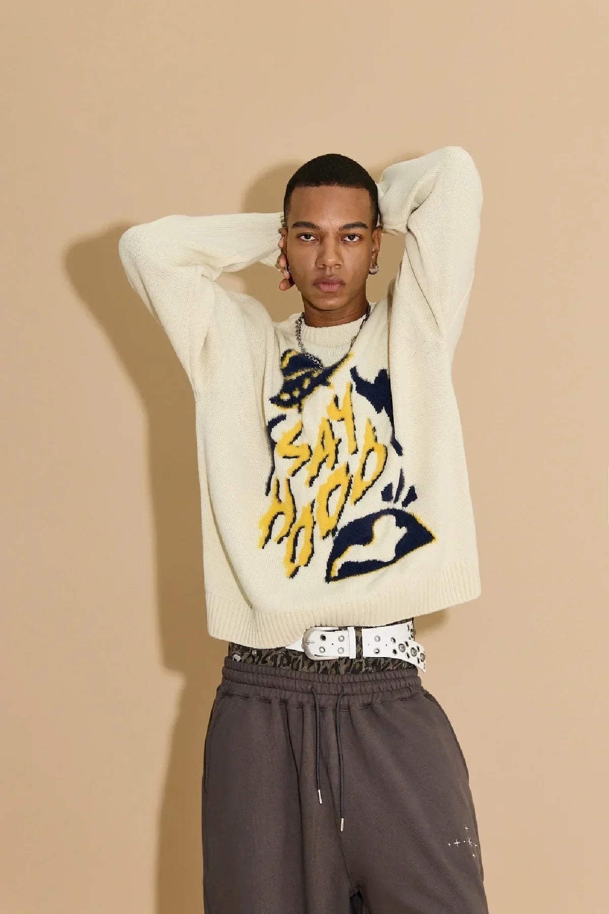 Young man wearing ivory knit sweater with bold yellow and blue Say Hood design and dark pants