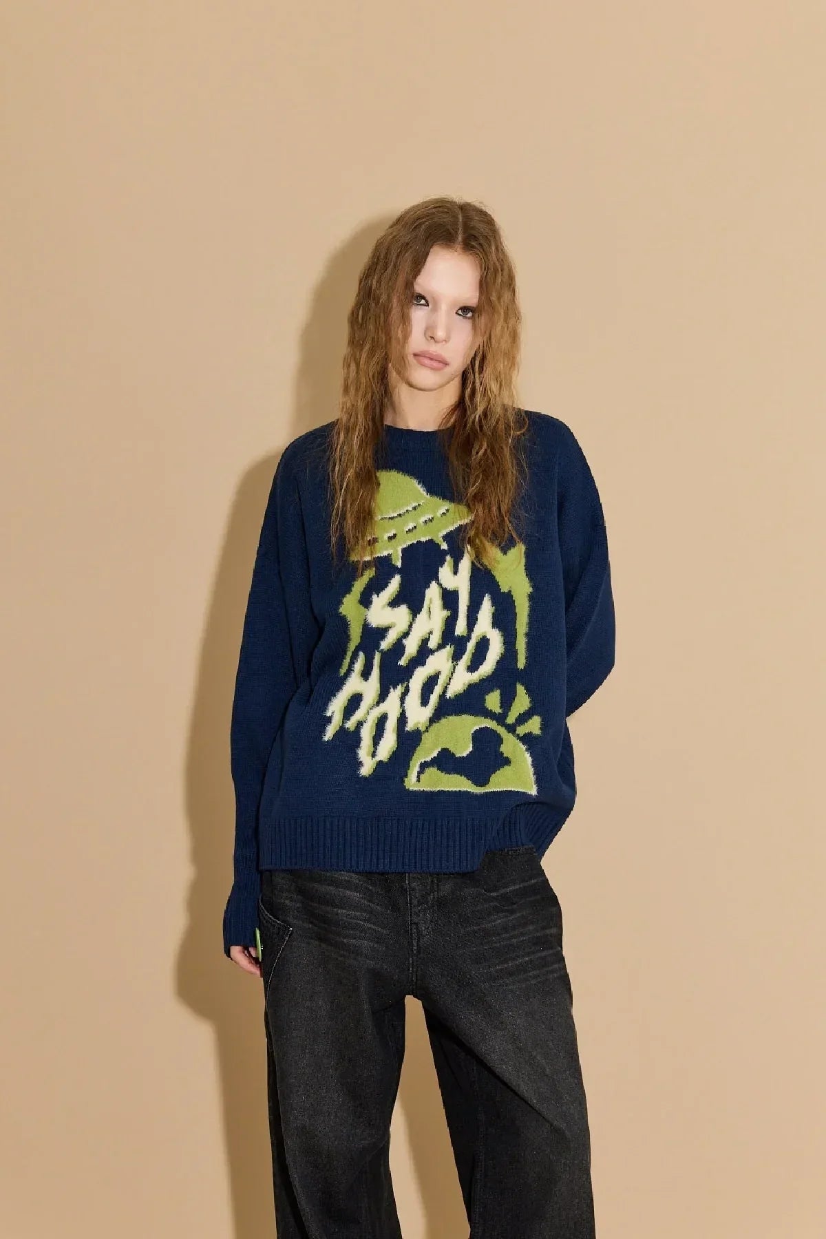 Model wearing navy blue knit sweater with green graphic and text, paired with black jeans against beige background